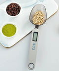Bilancia cucchiaio digitale Kiwi con display LCD che pesa legumi accanto a caffè e tè matcha, ideale per dosare ingredienti con precisione in cucina e pasticceria